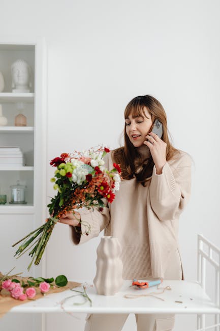 A woman with shoulder-length brown hair and bangs is holding a vibrant bouquet of fresh flowers, including red, white, pink, and green blooms, in her left hand while speaking on a mobile phone with he
