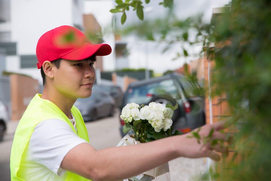 A young male florist wearing a red cap and a high-visibility yellow vest is arranging a bouquet of fresh white hydrangea flowers, which are wrapped in clear cellophane with a white ribbon. He is outdo