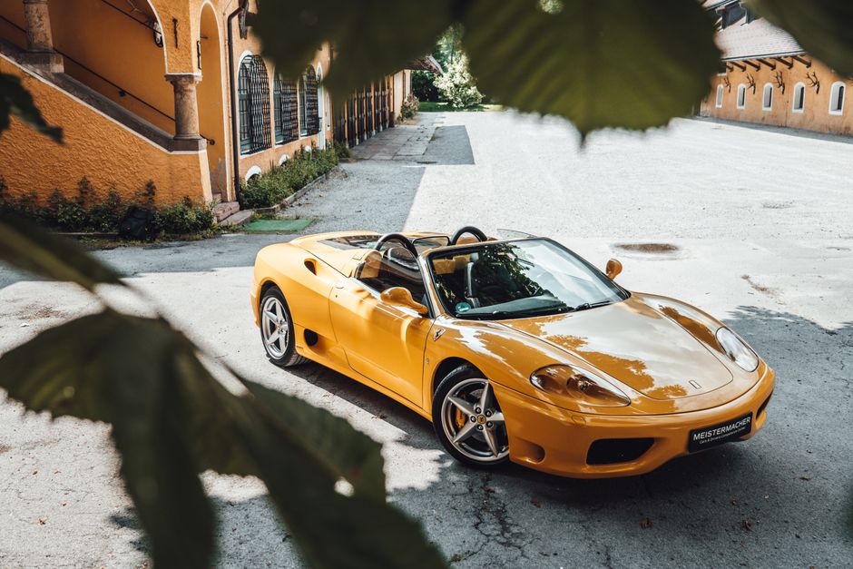 A bright yellow convertible sports car parked outdoors on a grey gravel surface, partially framed by green leaves in the foreground. The car has a sleek, aerodynamic design with a low profile, rounded