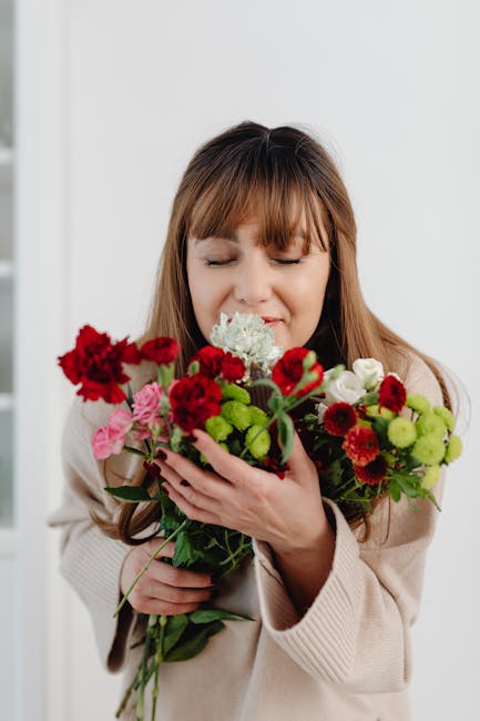 A woman with long brown hair and closed eyes holds a vibrant bouquet of fresh flowers close to her face. The bouquet features red, pink, and white blossoms, including small carnations, roses, and gree