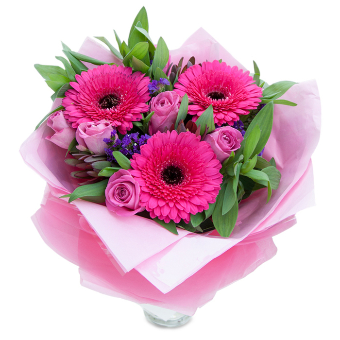 A carefully arranged posy that captures playful grace: three vivid fuchsia Gerbera daisies take centre stage, their many petals radiating outward and framing dark centres that give them structure and visual punch. Nestled between these statement blooms are several blush-to-lavender roses, each one tightly furled and promising a gentle unfurling that adds a sense of anticipation and tenderness. Flecks of purple statice are scattered through the bouquet like a fine sprinkling of confetti, creating delicate pockets of texture that contrast with the fuller rose forms. The supporting foliage is generous-lush salal leaves and slender eucalyptus stems-offering a crisp green backdrop and a faintly herbal scent that lifts the sweeter floral notes. The bouquet is swathed in a soft, matte pale pink tissue inner wrap, and a sheer, slightly crinkled fuchsia-pink cellophane layer over the top adds a whisper of shimmer and ties the whole pink-themed presentation together; the folds of the paper are neat, handcrafted, and intentionally arranged to hold the flowers snugly. Toward the bottom the rim of a clear glass vase is visible, suggesting the bouquet is designed for immediate display on a mantel, kitchen island, or bedside cabinet. In gentle daylight the combination of hot pink, dusty rose, and cool purple feels celebratory without being overpowering-perfect for marking a birthday, offering congratulations, or simply saying 'you're thought of'. I imagine this bouquet brightening a home near South Norwood Country Park or arriving as a thoughtful surprise on Croydon Road in the Elmers End neighbourhood.